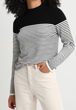Camisa de rayas blancas y negras de manga larga con un yugo negro texturizado; combinada con pantalones de color claro, mostrando un ajuste relajado.