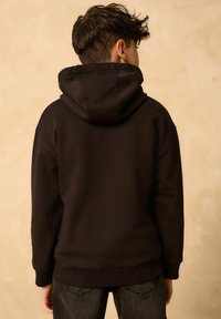 Dunkelbrauner Hoodie mit einer großen, verstellbaren Kapuze. Schlichtes Design, lange Ärmel, gerippte Bündchen und Saum. Kombiniert mit dunklen Jeans.
