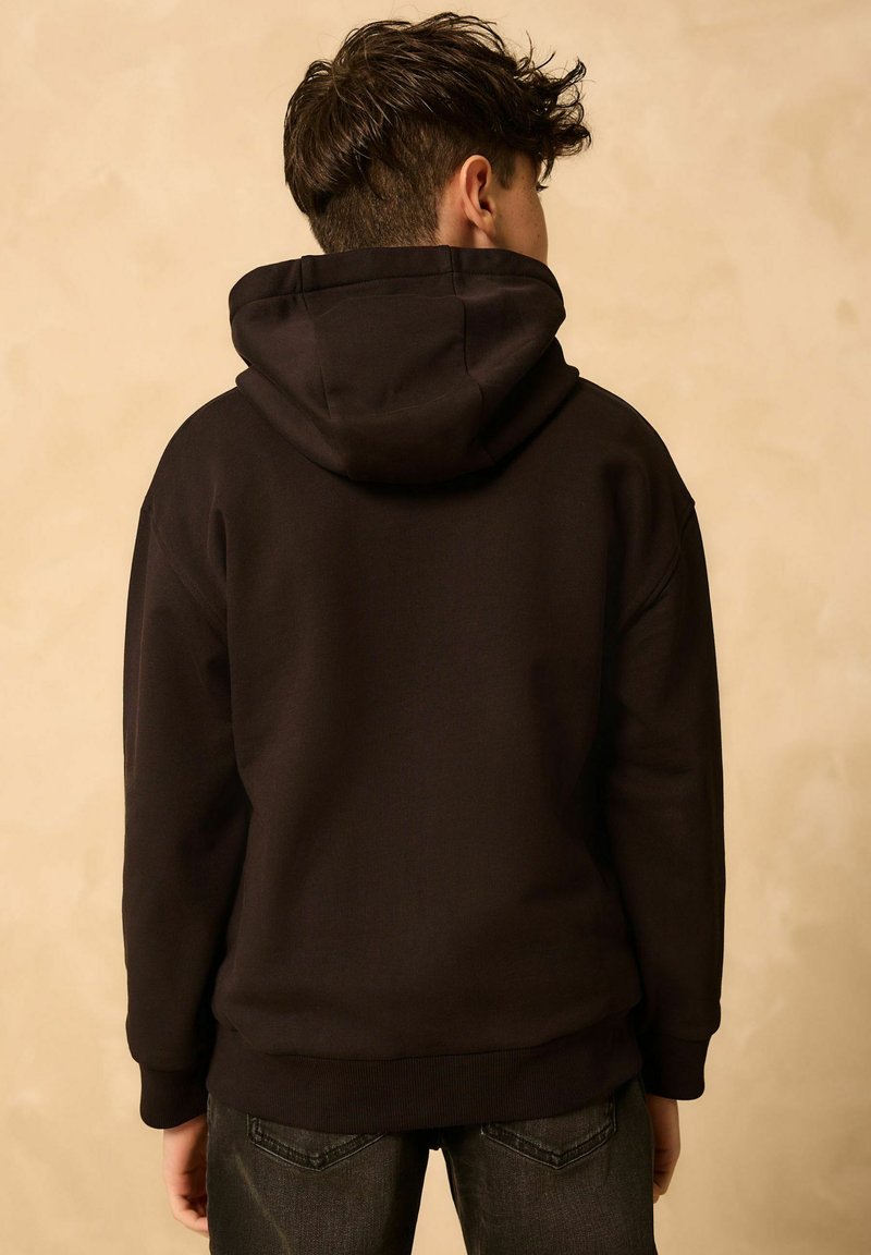 Dunkelbrauner Hoodie mit einer großen, verstellbaren Kapuze. Schlichtes Design, lange Ärmel, gerippte Bündchen und Saum. Kombiniert mit dunklen Jeans.