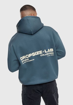 Mand iført en grønblå hættetrøje med teksten "DROPSIZE LAB COLLECTION" på ryggen, kombineret med sorte bukser, vendt væk mod en grå baggrund.