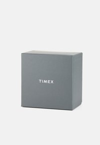 Boîte d'emballage grise avec une finition mate, arborant le logo "TIMEX" en texte blanc sur le devant. De forme carrée avec des lignes épurées.