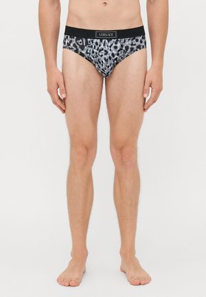 Mann steht barfuß und trägt schwarze und graue Leopardenmuster-Versace-Shorts vor einem schlichten weißen Hintergrund.