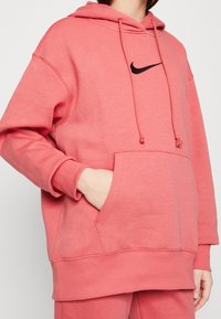 Kvinna som bär en lös korallrosa Nike-hoodie med en främre ficka och snören, hand placerad i fickan, kortad vid halsen.