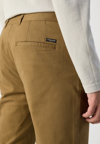 Bruine katoenen broeken met een zachte textuur, voorzien van een achterzak en een klein merklabel. De pijpen hebben een getailleerde pasvorm en strakke lijnen.