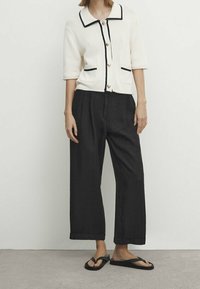 Kremowy, dziergany kardigan z czarnym obszyciem i guzikami, zestawiony z szerokimi czarnymi spodniami i czarnymi klapkami. Minimalistyczny styl, brak wzorów.