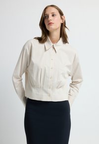 Przycięta, beżowa koszula na guziki z kołnierzykiem, długimi rękawami i plisowanymi detalami w talii, zestawiona z ciemną spódnicą.