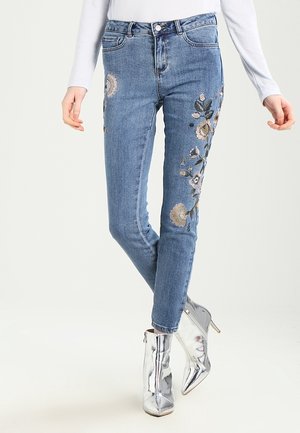 Person iført blå broderede blomstrede jeans kombineret med skinnende sølvfarvede høje ankelstøvler, stående med krydsede ben.