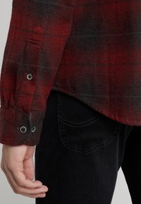 Camicia a quadri rossa e nera con tessuto testurizzato, dotata di polsini con bottone e orlo curvo, abbinata a jeans neri e una tasca posteriore.