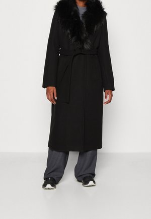 Person iført en lang sort frakke med pelskrave, grå vide bukser og sorte og hvide sneakers, står indendørs mod en hvid væg.