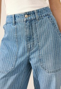 Pantalones vaqueros de denim azul claro con rayas blancas verticales, de cintura alta y con bolsillos delanteros. Presenta cierre con botón de metal y detalles de costura sutiles.