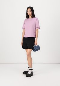 Lyse lilla kortermet skjorte med dekorative detaljer, parret med svarte shorts og svarte chunky sko, holder en liten blå veske.