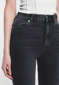 Korkeavyötäröiset mustat farkut, joissa on sileä pinta, hienovarainen haalistuma ja viiden taskun muotoilu. Varustettu metalliosilla ja erivärisillä ompeleilla.