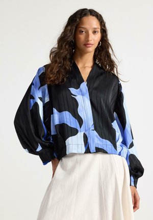 Nainen, jolla on pitkät kiharat hiukset, pukeutuneena löysään mustaan puseroon, jossa on sinisiä kukkakuvioita, ja valkoiseen pleiseerattuun hameeseen, seisomassa yksiväristä taustaa vasten.