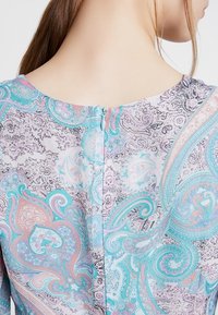 Femme avec de longs cheveux bruns attachés, portant une robe en pastel à motif paisley avec une fermeture éclair visible dans le dos, faisant face à l'arrière de l'appareil photo.