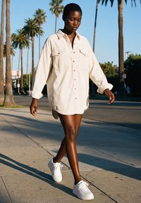 Sieviete pastaigājas pa ietvi, valkājot pārgrozušu bēšu pogājamu kreklu un baltas sporta kurpes, fonā redzamas palmas un skaidra debess.