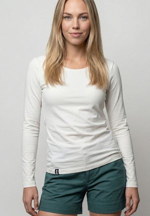 Junge Frau mit langen blonden Haaren, die ein weißes Langarmshirt und dunkelgrüne Shorts trägt, vor einem schlichten grauen Hintergrund stehend.