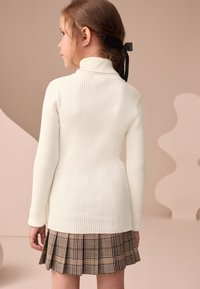 Pull à col roulé blanc côtelé associé à une jupe plissée à carreaux marron, présentant une texture subtile et une coupe ajustée. Ruban noir dans les cheveux.