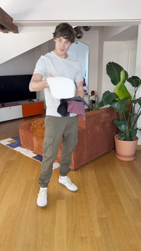 Modellen har en lys grå t-shirt og olivengrønne cargo bukser på, mens hun holder en hvid skjorte og to andre skjorter i mørke farver.