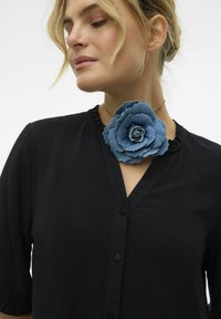 Černá košile s krátkým rukávem má kulatý výstřih a knoflíky, zdobená velkým broží ve tvaru květu z modrého denimu v oblasti límce.
