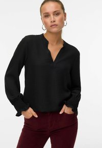 Donna che indossa una blouse nera a maniche lunghe con polsini arricciati e pantaloni color borgogna, in piedi con le mani infilate nelle tasche su uno sfondo bianco.
