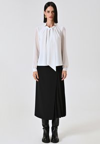 Blusa bianca con scollo drappeggiato e dettagli dorati, abbinata a una gonna nera plissettata. Stivali neri alla caviglia completano l'outfit.