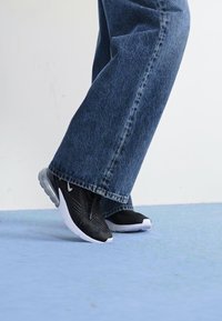 Sapatos atléticos pretos com um superior texturizado e seções de atacadores translúcido, combinados com calças de ganga azul largas sobre uma superfície azul clara.