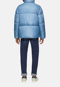 Lichtblauwe pufferjack met een hoge kraag, dikke padding en naden. Draagt het met donkere jeans en witte sneakers. Textuur van het materiaal zichtbaar.