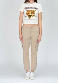 Besklippt vit t-shirt med en tigergrafik och text, tillsammans med beige cargo-byxor med sidofickor, bärs med vita sneakers.