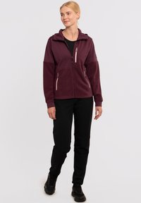 Veste en polaire bordeaux avec fermeture éclair à l'avant, capuche et deux poches latérales. Associée à un pantalon noir et des chaussures noires, se tenant devant un fond uni.