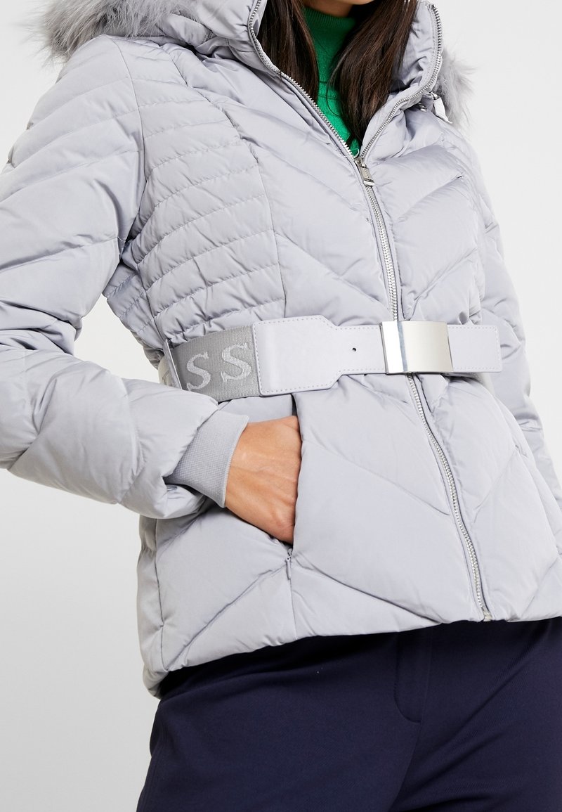 Femme portant une veste matelassée gris clair avec capuche, large ceinture assortie, et pull vert, main dans la poche de la veste, pantalon bleu marine visible.