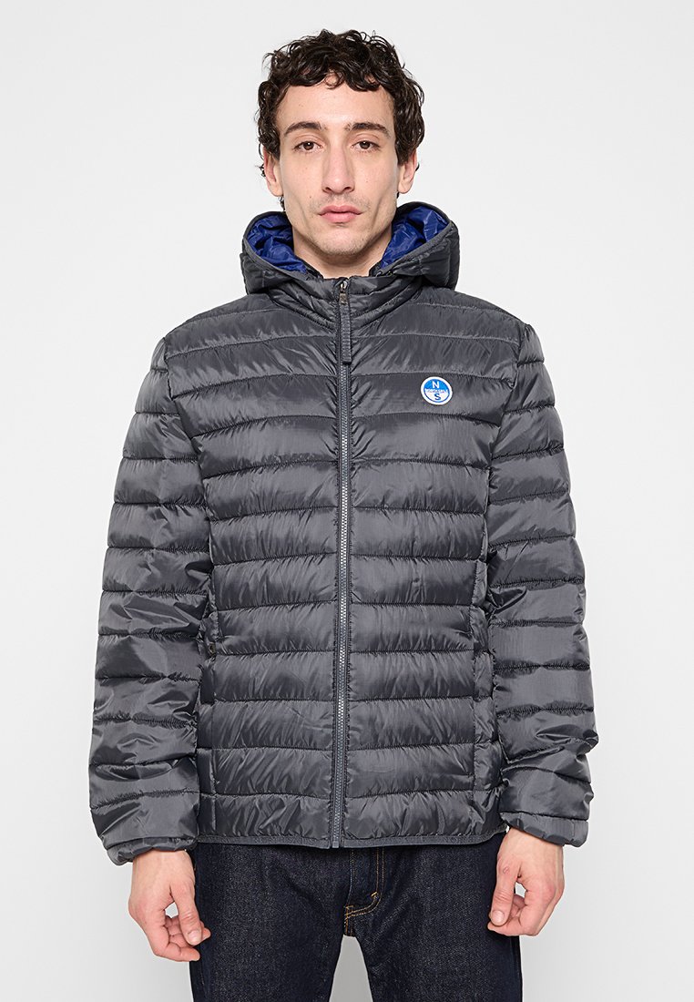 Giacca grigia imbottita con chiusura a zip, dotata di foderatura blu nel cappuccio e una patch con logo sul petto; texture liscia e cuciture orizzontali.