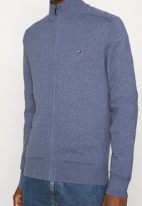Maglione azzurro chiaro, con zip e collo alto, realizzato in un tessuto a maglia testurizzato, con un piccolo logo sul petto.