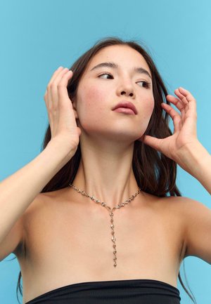 Collana dorata con elementi a forma di diamante alternati, dotata di un pendente centrale a goccia, su uno sfondo azzurro chiaro.