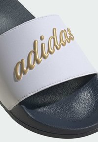 Białe klapki z fakturowaną granatową podeszwą, z uniesionym złotym logo "adidas" na pasku i widocznymi detalami szycia.