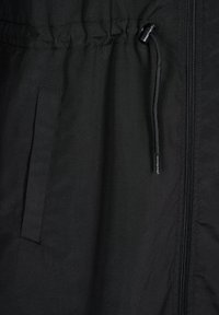 Schwarze Jacke aus strukturiertem Stoff, mit elastischem Bund, Reißverschluss und seitlicher Tasche. Enthält einen Kordelzug für eine verstellbare Passform.