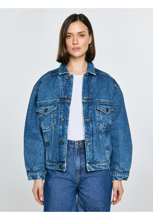 Sieva ar taisniem tumšiem matiem, kas valkā pārizmēra zilas džinsa jaku, baltu kreklu un zilas augsta vidukļa džinsas, stāvot pret vienkāršu fonu.