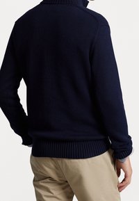 Marineblauer Strickpullover mit hohem Kragen und gerippten Bündchen, kombiniert mit beigen Hosen, der ein strukturiertes, gemütliches Design zeigt.