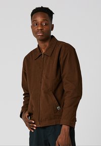 Καφέ τζάκετ με φερμουάρ και γιακ ά, με λεία υφή και πλαϊνές τσέπες. Μινιμαλιστικός σχεδιασμός χωρίς εμφανή μοτίβα ή αξεσουάρ.