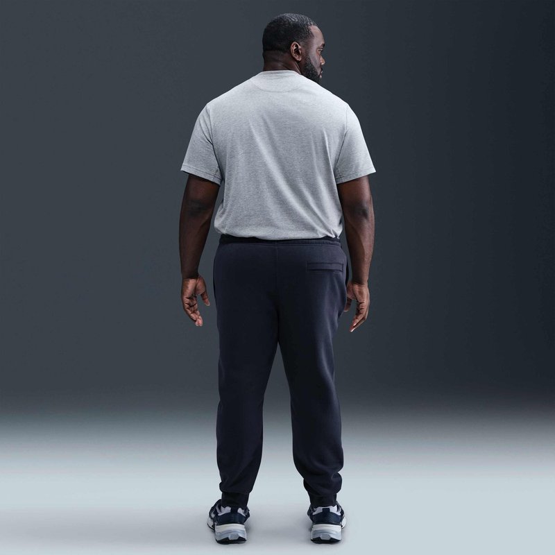 Uomo in piedi di spalle alla fotocamera, indossa una maglietta grigia, pantaloni scuri e sneaker, su uno sfondo scuro e semplice.