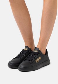 Svarta lädersneakers med en slät yta, rund tå, platta svarta sulor och guldiga logodetaljer på tungan och sidan.