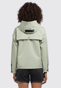 Lekka zielona wodoodporna kurtka z wysokim kołnierzem, przednią klapą i logo. Posiada długie rękawy oraz regulowane mankiety. Noszona na czarnych szortach.