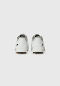 Baskets de sport blanches au texture lisse, avec des accents gris et des semelles noires équipées de pointes de traction orange. Comprend une poignée au talon.