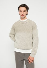 Πλεκτό πουλόβερ σε ανοιχτό μπεζ με υφή, λαιμόκοψη στρογγυλή και άνετη εφαρμογή, φορεμένο πάνω από λευκό πουκάμισο με γκρι παντελόνι.