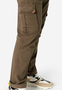 Cargo-Hosen aus braunem Stoff mit Karomuster, ausgestattet mit Seitentaschen und einem umgekrempelten Saum, kombiniert mit beige-weißen Sportschuhen.