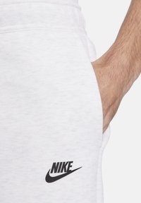 Balti sporta bikses, kas izgatavotas no mīksta kokvilnas auduma, ar melnu Nike logotipu uz kreisā kājas un sānu kabatu. Gluda tekstūra.