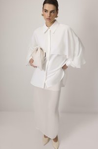 Hvid oversized skjorte med knaplukning og buet kant, parret med en gennemsigtig hvid nederdel. Holder en beige clutch. Lyse spidse hæle.