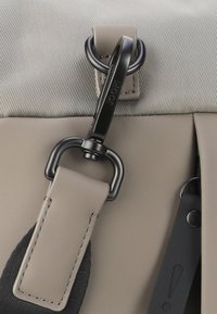 Bežová taška s texturovanou tkaninou, s černými kovovými prvky, klipem a černým uchycením popruhu s visačkou zobrazující vykřičník.