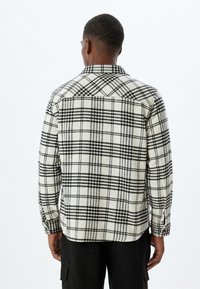 Camisa a cuadros en beige y negro, hecha de una tela suave. Presenta mangas largas, una tapeta de botones y un cuello clásico con un patrón de cuadros pronunciado.