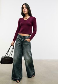Femme portant un pull bourgogne court, un jean noir délavé à jambes larges, des talons ouverts noirs, tenant un sac à main noir, debout sur un sol gris.
