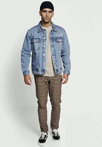 Casaco de ganga com um tom azul claro, dois bolsos no peito e detalhe em corrente. Combinado com calças castanhas estampadas e ténis pretos.